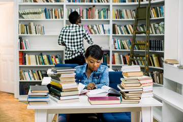 African american young woman reading textbook sitting at the table on the background of bookshelf.Concentrated dark sinned student spending leisure time in library
