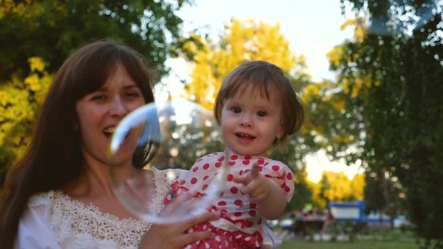 Happy mom and daughter catch big soap bubbles in hot summer park and laugh. Nanny with little girl playing for walk and smiling.