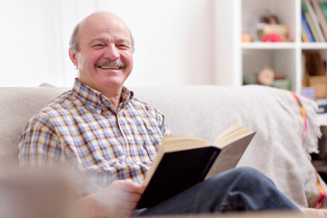 Naklejka premium Senior man reading a book on his sofa