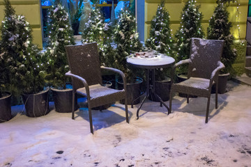 Fototapeta na wymiar The table and two armchairs are covered with snow on the table is a garland. On the background can see the fir trees.
