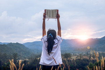 young woman holding and lift up bible to sky outdoors with light sunset background,christian...
