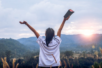 young woman holding bible and lift up hand with light sunset background,christian concept.