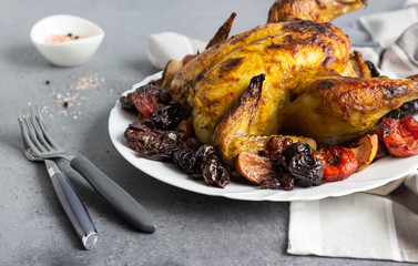 Tasty food: whole roasted chicken with dried fruits (raisin, apricot and plum) and apples on a white plate. Grey stone background. Copy space. 
