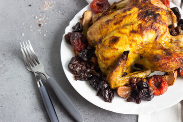 Tasty food: whole roasted chicken with dried fruits (raisin, apricot and plum) and apples on a white plate. Grey stone background. Copy space. 