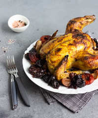 Tasty food: whole roasted chicken with dried fruits (raisin, apricot and plum) and apples on a white plate. Grey stone background. Copy space. 