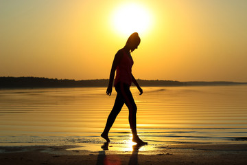 Fototapeta na wymiar Silhouette of a young beautiful girl walking along the seashore during orange sunset