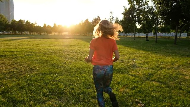 Sports woman doing jogging on the grass on sunset in a city park, slow motion