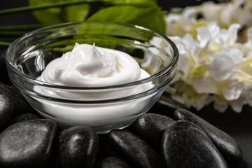 Close up of natural skin cream in glass bowl on spa like rock base white flowers in background.