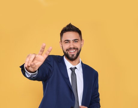 Happy young businessman showing victory gesture on color background