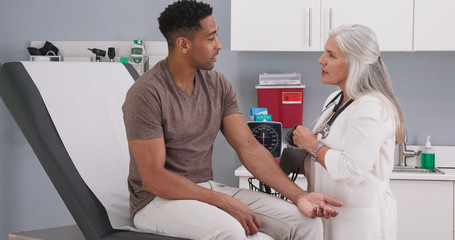 Handsome african-american patient seated in medical room having his blood pressure checked by...