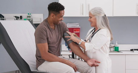 Handsome african-american patient seated in medical room having his blood pressure checked by senior doctor. Mature white female physician checking male patients blood pressure in health clinic - Powered by Adobe