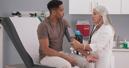 Senior caucasian doctor using stethoscope and gauge to measure patients blood pressure. Young black...