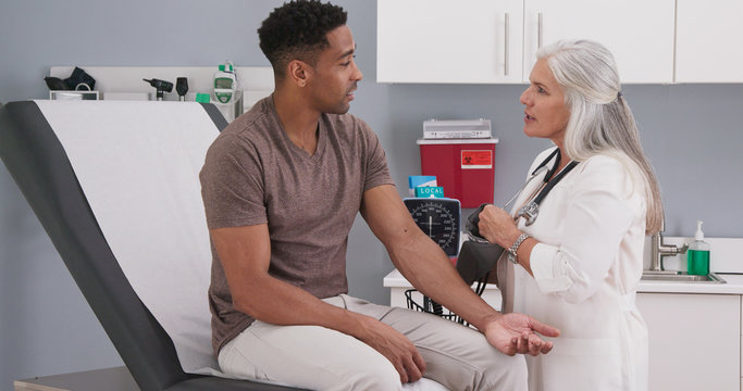 Handsome african-american patient seated in medical room having his blood pressure checked by senior doctor. Mature white female physician checking male patients blood pressure in health clinic