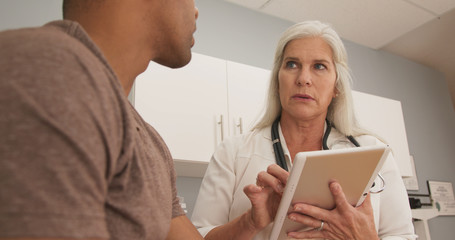 Closeup of mature female doctor using tablet computer to take notes of patient. Male...