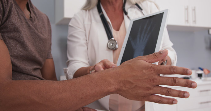 Closeup Of Black Patient Holding Out Injured Hand And Wrist While Doctor Shows Xray On Tablet. Close Up Of Doctor Explaining X-ray And Cause Of Pain To Male Patient