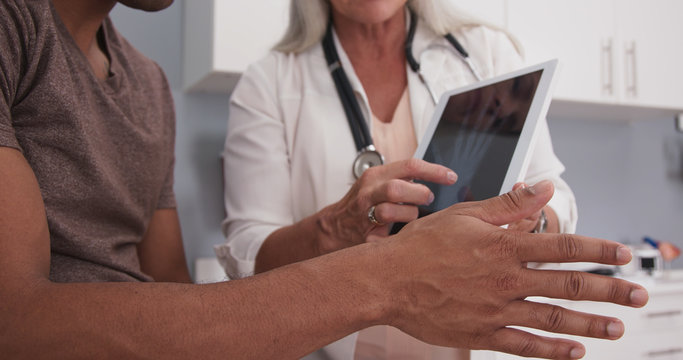 Closeup Of Black Patient Holding Out Injured Hand And Wrist While Doctor Shows Xray On Tablet. Close Up Of Doctor Explaining X-ray And Cause Of Pain To Male Patient