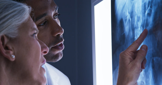 Close view of young male doctor and senior colleague looking at x-ray of patients spine. Close up of millennial black doctor consulting with senior colleague over x ray of spinal bones - Powered by Adobe