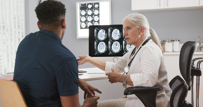 Young african-american patient meeting with senior medical doctor about concussion. Elderly female doctor showing ct scans of patients brain - Powered by Adobe