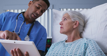 Male millennial black nurse using tablet computer to show senior woman images of her surgery procedure. Young RN and elderly patient looking at electronic pad