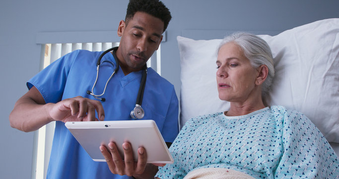 Young african-american RN and senior patient looking at tablet computer at hospital. Male registered nurse showing ill elderly woman electronic pad