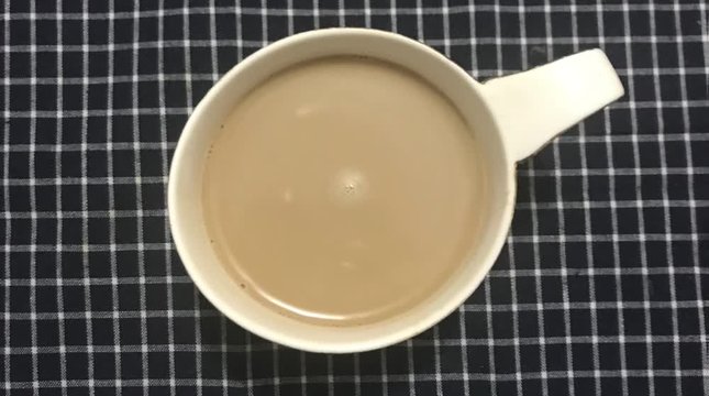 Cinemagraph - White cup of stirred coffee on a table with a checkered tablecloth. Top view. Closeup