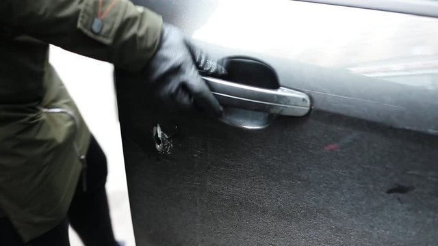 Woman wearing jacket and black leather gloves and holding thin metallic stick with curved tip is aggressively making big hole in surface of car door at street in day light and opens it. Robbery.