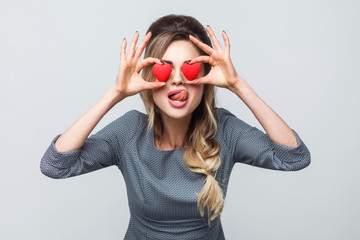 Loving eyes. Beautiful sexy caucasian young woman holding two valentine hearts in front of her eyes like glasses with sensual tongue in grey background. Copy space, indoor, studio shot, isolated