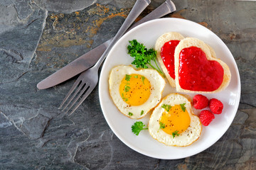Valentines Day brunch concept with heart shaped eggs and toast with jam over a dark stone background. Top view.