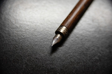 Macro shot with shallow depth of field focusing on metal nib with fine iridium point and engraved metal grip with cylindrical walnut wood body set on textured black table with reflective light halo.