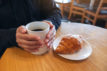 A woman holding coffee cup with a piece of croissant in a white plate on wooden table in cafe