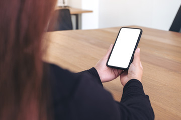 Mockup image of a woman holding black mobile phone with blank white screen on wooden table in office