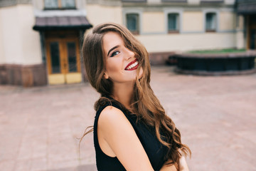 Portrait of pretty girl with long curly hair posing to camera on street on old building background. She wears black dress, vinous lips. Hair covers half her face, she is smiling.