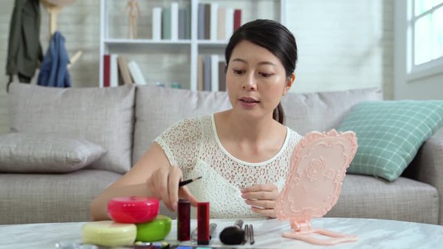 Female beauty asian vlogger showing pink lipstick and brush teaching makeup to video camera online sitting in living room indoors. Live stream young people selling cosmetic. like and share teaching.