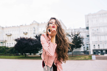 Fototapeta premium Attractive girl with long hairstyle having fun in city. She has pink jacket, sending a kiss with vinous lips to camera.