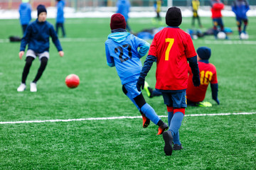 Obraz premium Boys in red and blue sportswear plays soccer on green grass field. Youth football game. Children sport competition, kids plays outdoor, winter activities, training