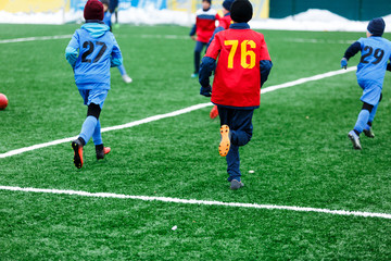 Obraz premium Boys in red and blue sportswear plays soccer on green grass field. Youth football game. Children sport competition, kids plays outdoor, winter activities, training
