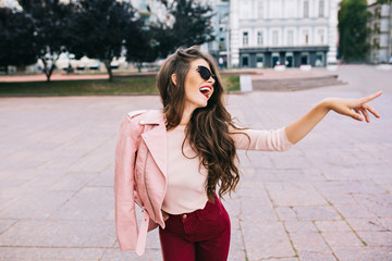 Fototapeta premium Elegant girl with long hairstyle in vinous pants having fun in city. She has pink jacket on shoulder, showing to side.