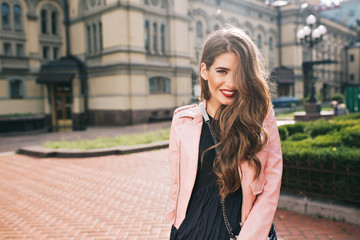 Fototapeta premium Portrait of young girl with long curly hair and red lips posing on steer on building background. She wears black dress, pink jacket. Hair covers half her face and she is smiling to camera.