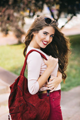 Portrait of cute girl with long hair and vinous lips smiling to camera in city park. She wears marsala bag, pants and white shirt.