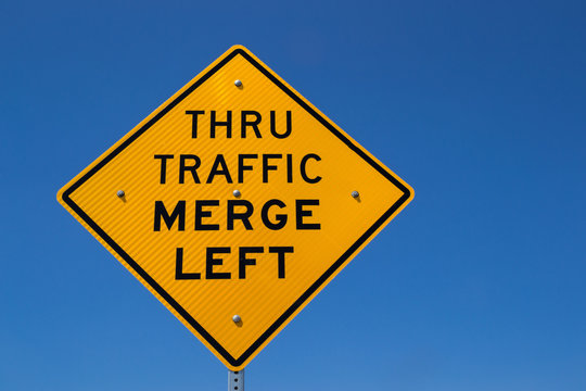 The "Thru Traffic Merge Left" street sign with brilliant blue skies in the background.