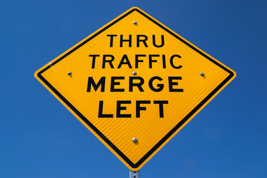 The "Thru Traffic Merge Left" street sign with brilliant blue skies in the background.