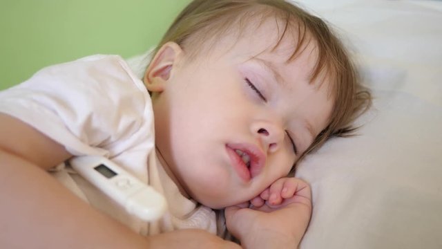 Small child sleeps in hospital ward on white bedding and measures temperature with thermometer. Treatment of children in hospital. Sick baby improves his health in hospital.