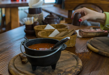 Russian soup, borscht, in a cast iron pot on the background of a woman hand, on a wooden table in a stylish restaurant, in the old Russian style