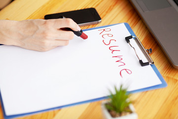 Job search. Female hand writes resume with lipstick on white sheet of paper. Wooden office desk with laptop, smartphone and supplies