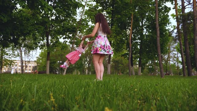 Young mother turns her little daughter around and laughs. Happy mother and child having fun in park together and smiling. Nanny walks with kid in city.