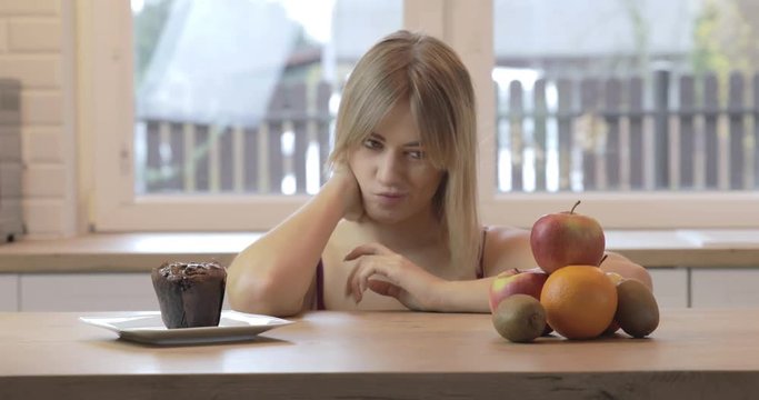 Close-up of a conflicted woman with a dilemma. She makes silly faces while trying to decide between healthy food and junk food plates that are on the table in front of her.