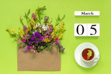 Wooden cubes calendar March 5. Cup of herbs tea, kraft envelope with multi colored flowers on green background. Concept hello spring Creative Top view Flat lay