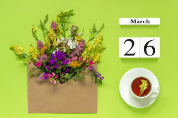 Wooden cubes calendar March 26. Cup of herbs tea, kraft envelope with multi colored flowers on green background. Concept hello spring Creative Top view Flat lay