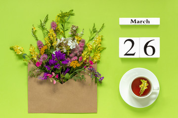 Wooden cubes calendar March 26. Cup of herbs tea, kraft envelope with multi colored flowers on...