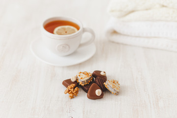 Cup of hot tea with lemon, saucer, chocolate candy with nuts and knitted sweaters on white wooden table, selective focus.